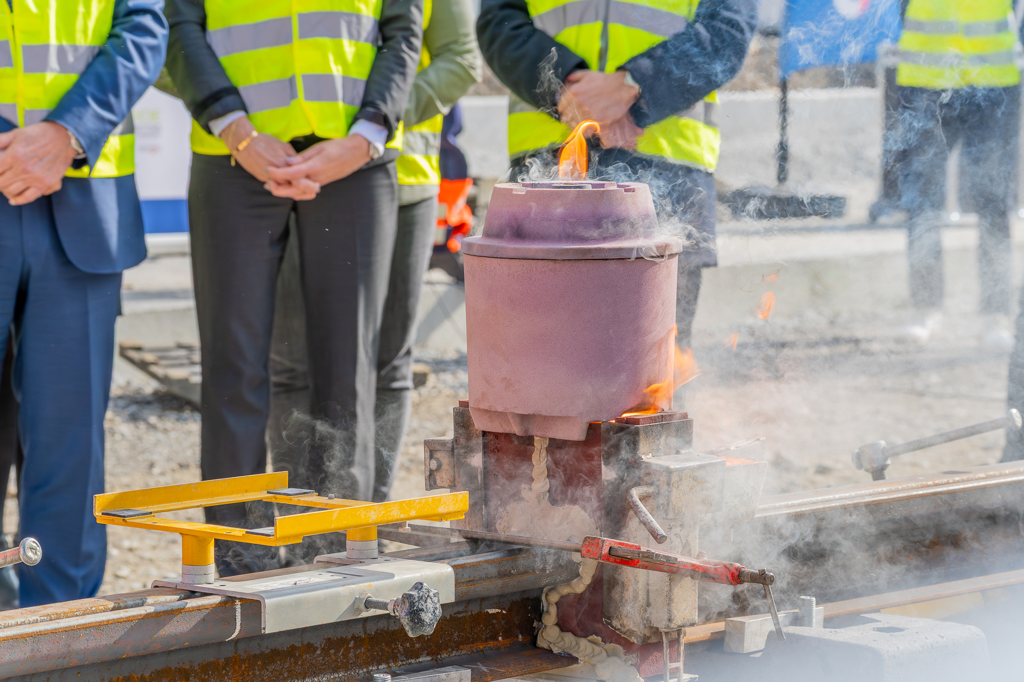 Opération de soudure aluminothermique sur des rails de tramway. Au premier plan, on voit un creuset contenant un mélange incandescent en fusion qui dégage de la fumée et des flammes orangées. Le dispositif est fixé aux rails qui seront soudés. En arrière-plan, plusieurs personnes portant des gilets de sécurité jaune fluorescent observent l'opération, certainement des officiels et techniciens présents pour cette cérémonie symbolique.